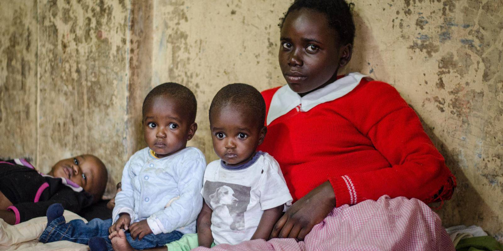 A young mother sitting with three small children against a bare wall.